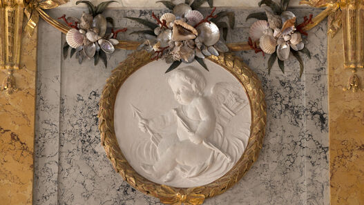 Schloss Rheinsberg, Flachrelief mit Puttenszene an der oberen Westwand des Muschelsaals © SPSG / Leo Seidel Schloss Rheinsberg, Muschelsaal, Dekorationselement mit dem lorbeerkranzartigem gerahmtem ovalem Flachrelief mit Puttenszene (Amor) an der oberen Westwand rechts neben der Tür im Muschelsaal