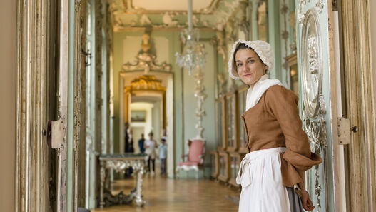 Eine als Kammerzofe verkleidete junge Frau steht an einer geöffneten Tür, hinter ihr ist Schlossraum zu sehen