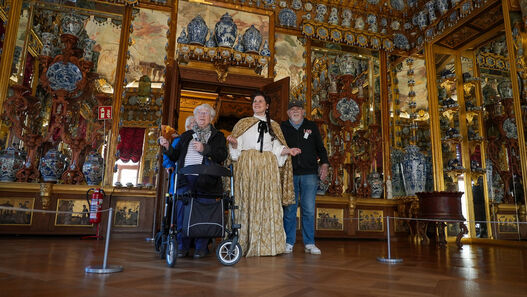 Führung der Malteser durch das Schloss Charlottenburg für Menschen mit Demenz, zu sehen sind eine Darstellerin als Königin Sophie Charlotte mit einem älteren Mann mit grauem Bart und Basecap sowie einer älteren Frau mit Rollator im Porzellankabinett
