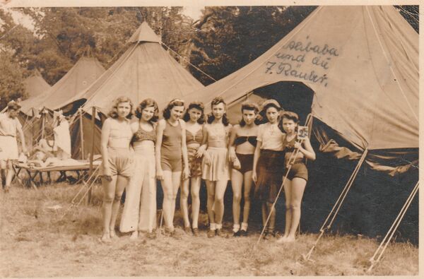 Acht junge Mädchen stehen sommerlich bekleidet vor einem elt. darüber steht handgeschrieben „Alibaba und die 7. Räuber“, das Foto entstand 1948