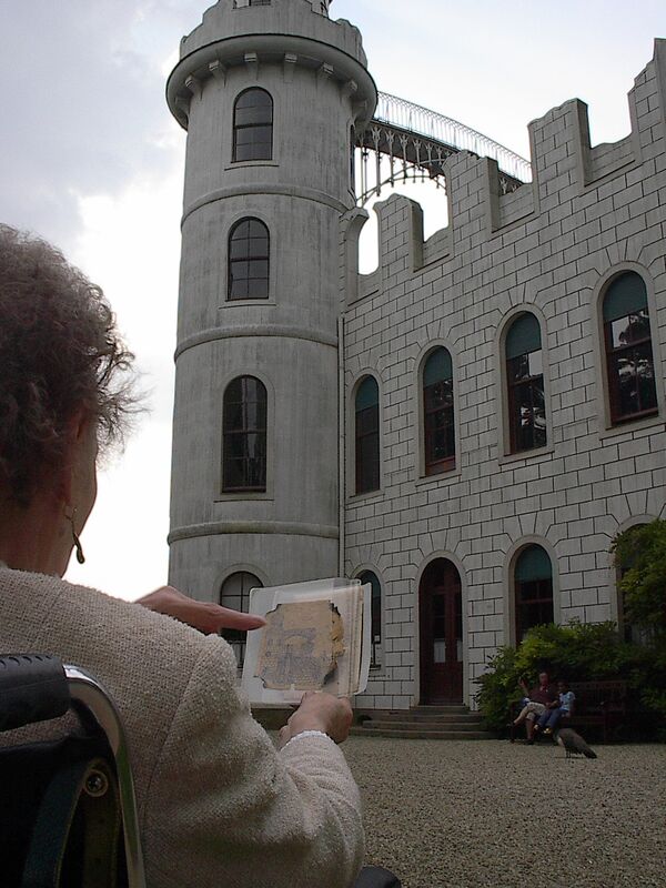 Eine ältere Dame im Rollstuhl sitzend von hinten fotografiert; in der Hnad hält sie eine Zeichnung des Pfaueninselschlosses, im Hintergrund das Pfaueninselschloss