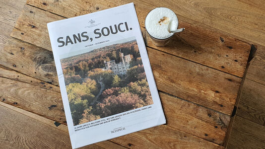 SANS, SOUCI. Das Magazin der Stiftung Preußische Schlösser und Gärten Berlin-Brandenburg, Ausgabe 4.2025 auf einem Tisch, daneben ein Kaffeeglas mit Löffel