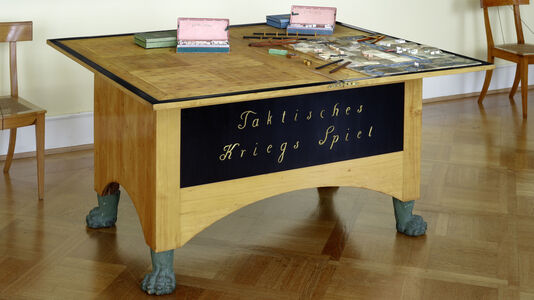 Domänen- und Kriegsrat Georg Leopold Baron von Reisswitz, Kommode „Taktisches Kriegs Spiel" für Friedrich Wilhelm III., 1812.  Das später weiter entwickelte Kriegsspiel befindet sich heute im Neuen Flügel des Schlosses Charlottenburg, im Raum 314. Was für die Besuchenden wie eine schlichte Kommode anmutet, verbirgt in seinem Inneren unbegrenzte Möglichkeiten an Kriegssimulationen.