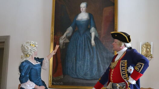 Kostümführung mit Manuela und Mike Sprenger, Manuela Sprenger trägt ein Kleid, das dem von Sophia Dorothea auf dem Bild hinter ihr zu sehen ist