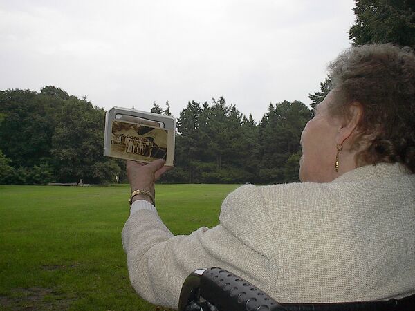 Eine alte Dame von hinten fotografiert, die ein Schwarz-Weiß-Foto vor sich in die Höhe hält, im Hiintergrund eine Wiese und Bäume